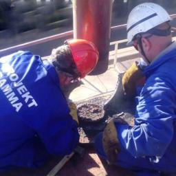 Projekt Gamma Sp. zoo - Dwóch pracowników w kombinezonach z napisem 'PROJEKT GAMMA' w kaskach, wykonuje prace spawalnicze na konstrukcji stalowej, widoczne iskry i narzędzia.