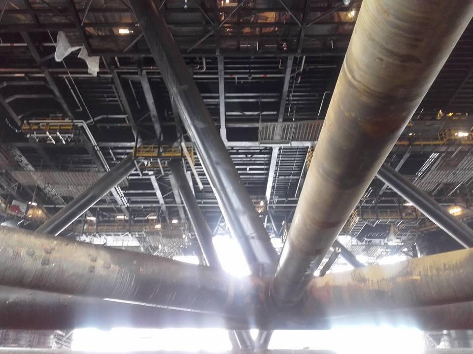 Wnętrze konstrukcji stalowej hali z widocznymi rurami nośnymi i metalową siatką sufitową, oświetlone od dołu silnym światłem.