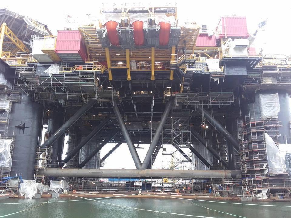 Rozbudowana platforma wiertnicza na wodzie, otoczona rusztowaniami i elementami konstrukcyjnymi, widok z dołu w pochmurny dzień.