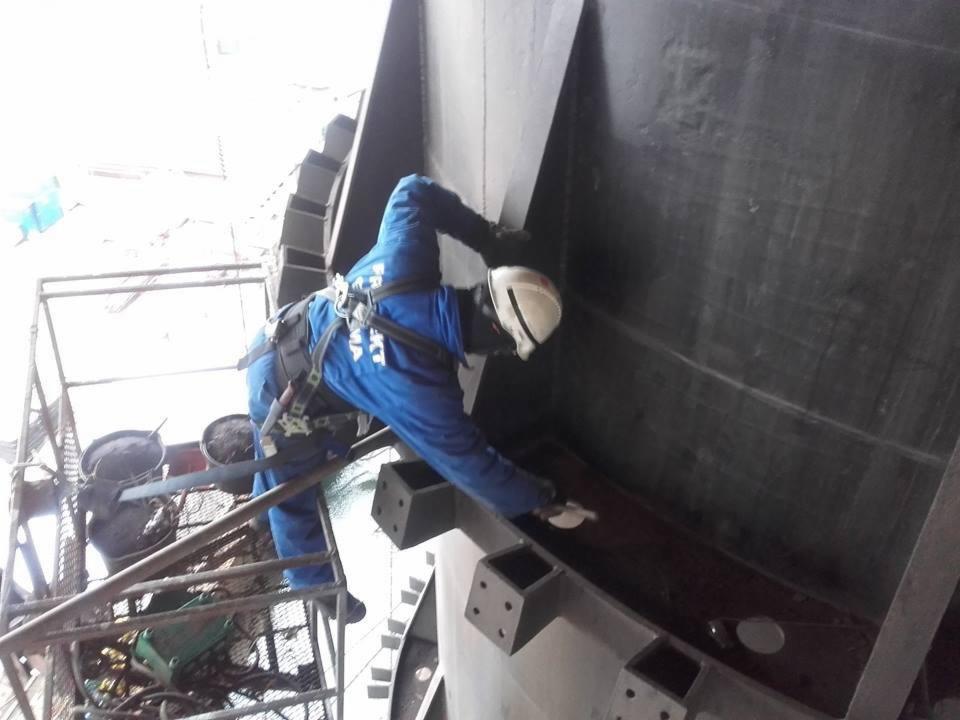 Pracownik w kombinezonie i kasku, zabezpieczony linami, maluje wewnętrzną powierzchnię stalowej konstrukcji hali, widoczne rusztowanie i wiadro z farbą.