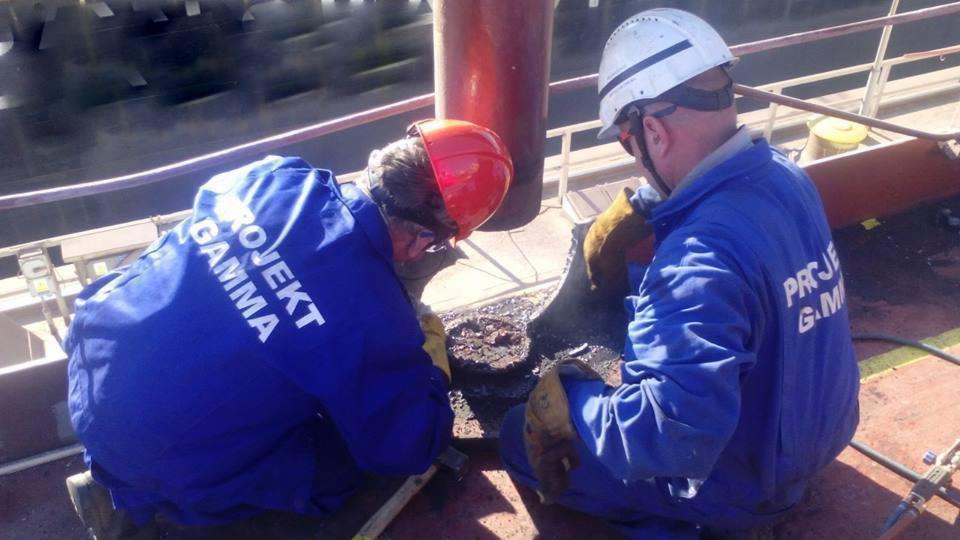 Dwóch pracowników w kombinezonach z napisem 'PROJEKT GAMMA' w kaskach, wykonuje prace spawalnicze na konstrukcji stalowej, widoczne iskry i narzędzia.