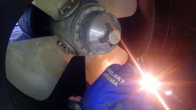 Spawanie metalowej konstrukcji w hali, iskry i niebieska odzież ochronna z logo Projekt Gamma.