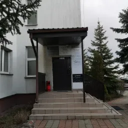 Wejście do budynku z szyldami firm, w tym kancelarii adwokackiej i archiwum, z widoczną nazwą adwokata Anna Dyka, schody z brązowymi płytkami, zieleń w tle.