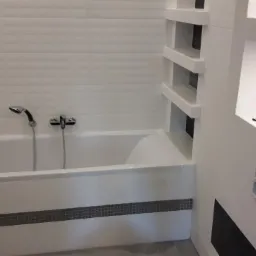 Nowoczesna, biała łazienka po remoncie: wanna obudowana płytkami z dekoracyjnym pasem mozaiki, wnęki ścienne z p&oacute;łkami i mozaiką, bateria wannowa z deszczownicą.