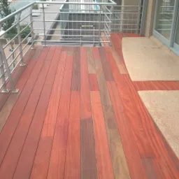 Wykończony balkon z drewnianą podłogą ułożoną z desek o różnych odcieniach czerwieni i brązu, z metalową balustradą i fragmentem elewacji budynku z przesuwnymi drzwiami balkonowymi.