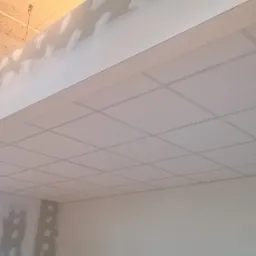 Sufit podwieszany z płyt gipsowo-kartonowych z widocznymi śladami szpachlowania na połączeniach, oświetlenie sufitowe w tle, widok z dołu na fragment pomieszczenia w trakcie prac wykończeniowych.