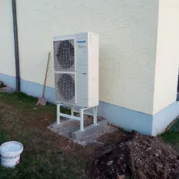 Biała zewnętrzna jednostka klimatyzacji Panasonic Inverter na metalowej konstrukcji, zamontowana przy elewacji budynku z widoczną łopatą opartą o ścianę i stertą ziemi w pobliżu.