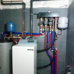 Kotłownia z kotłem gazowym Pulsatoire, zasobnikiem ciepłej wody, naczyniami przeponowymi i instalacją hydrauliczną z rur miedzianych i z tworzyw sztucznych w pomieszczeniu wyłożonym szarą glazurą.