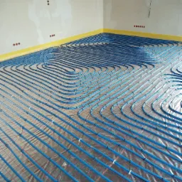 Niebieskie rury układane w pętle na siatce grzewczej podłoża, widoczne punkty mocowania, żółta taśma przy ścianie, surowe ściany z puszkami elektrycznymi.