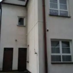 Biały budynek dwukondygnacyjny z widoczną kamerą monitoringu na elewacji, brązowe drzwi wejściowe i okna z białymi ramami.