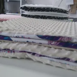 Stos uszytych kocyków dziecięcych z miękkiej tkaniny minky w kolorze kremowym, wykończonych bawełną z motywem piór, leżący na białym stole w szwalni.