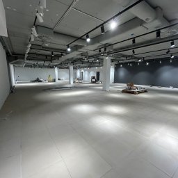 Usługi Sebastian Walczak - Przestronne wnętrze sklepu w trakcie wykańczania, widoczne białe płytki na podłodze, surowy sufit z licznymi reflektorami, białe ściany i ciemna ściana w tle, materiały budowlane na paletach...