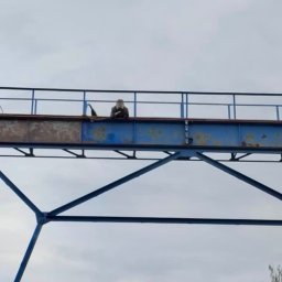 Usługi Sebastian Walczak - Pracownik w kombinezonie na wysokości, na konstrukcji stalowej zardzewiałej w trakcie prac konserwacyjnych, widoczna drabina.