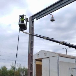 Usługi Sebastian Walczak - Pracownik w podnośniku koszowym czyści metalową konstrukcję na zewnątrz, widoczne koła kolejowe, drabina i biały namiot w tle, pochmurne niebo.