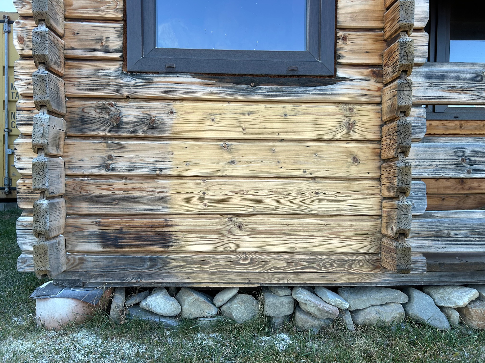 Fragment ściany drewnianej chaty z oknem, widoczne ślady zabrudzeń i przebarwień na drewnie, kamienna podmurówka.