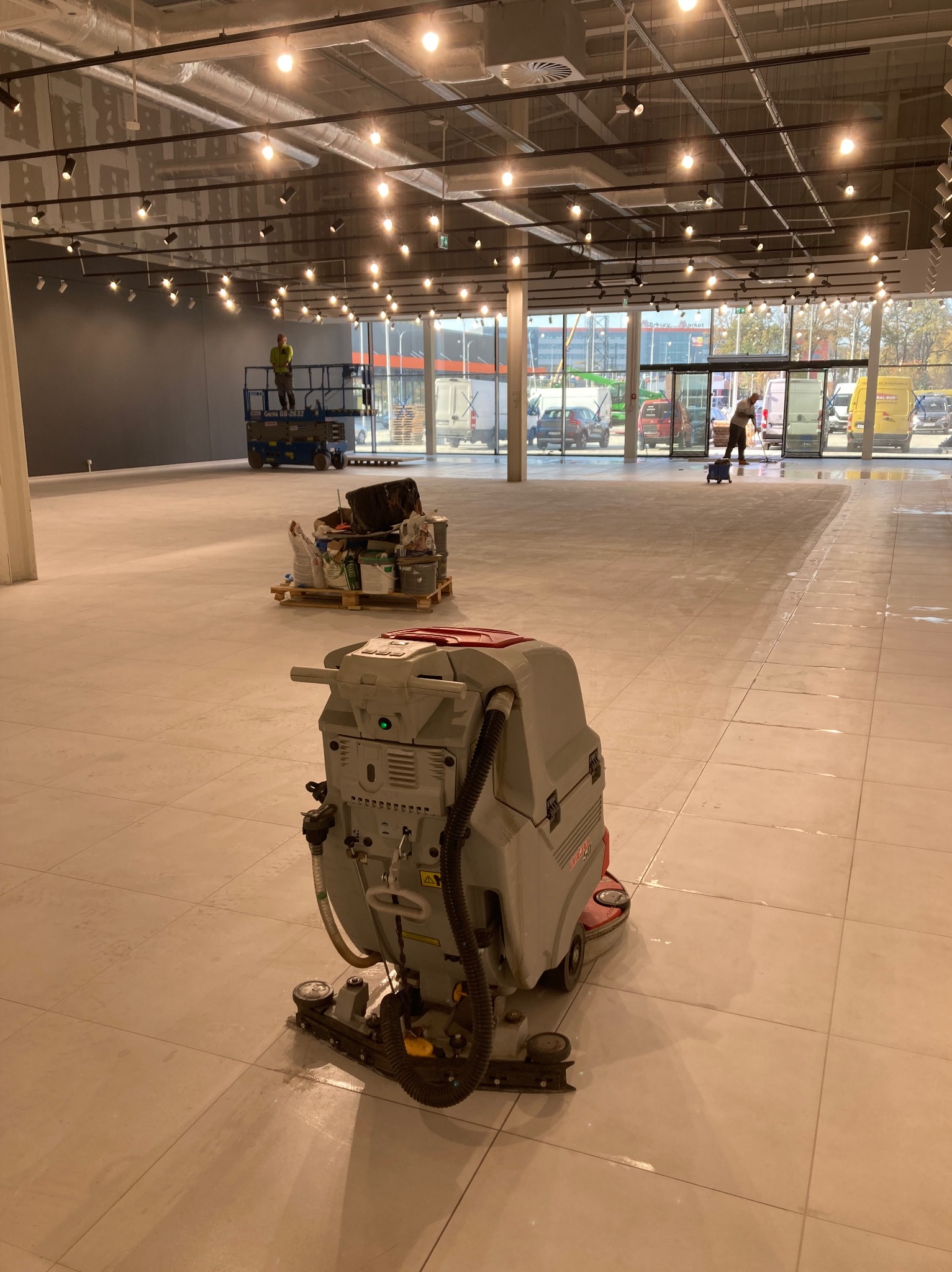 Duża powierzchnia hali w trakcie czyszczenia posadzki przy użyciu automatycznej szorowarki, widoczny pracownik myjący podłogę mopem w oddali oraz podnośnik koszowy z operatorem przy ścianie.