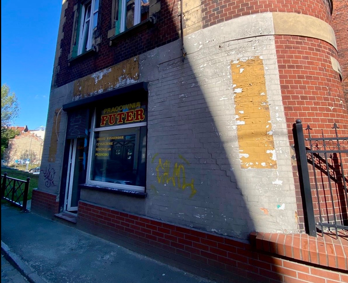 Zewnętrzna ściana budynku z cegły i szarego tynku, z widocznymi śladami odpadającej farby i graffiti. Na ścianie okno z szyldem 'Pracownia Futer' oraz informacją o usługach kuśnierskich...
