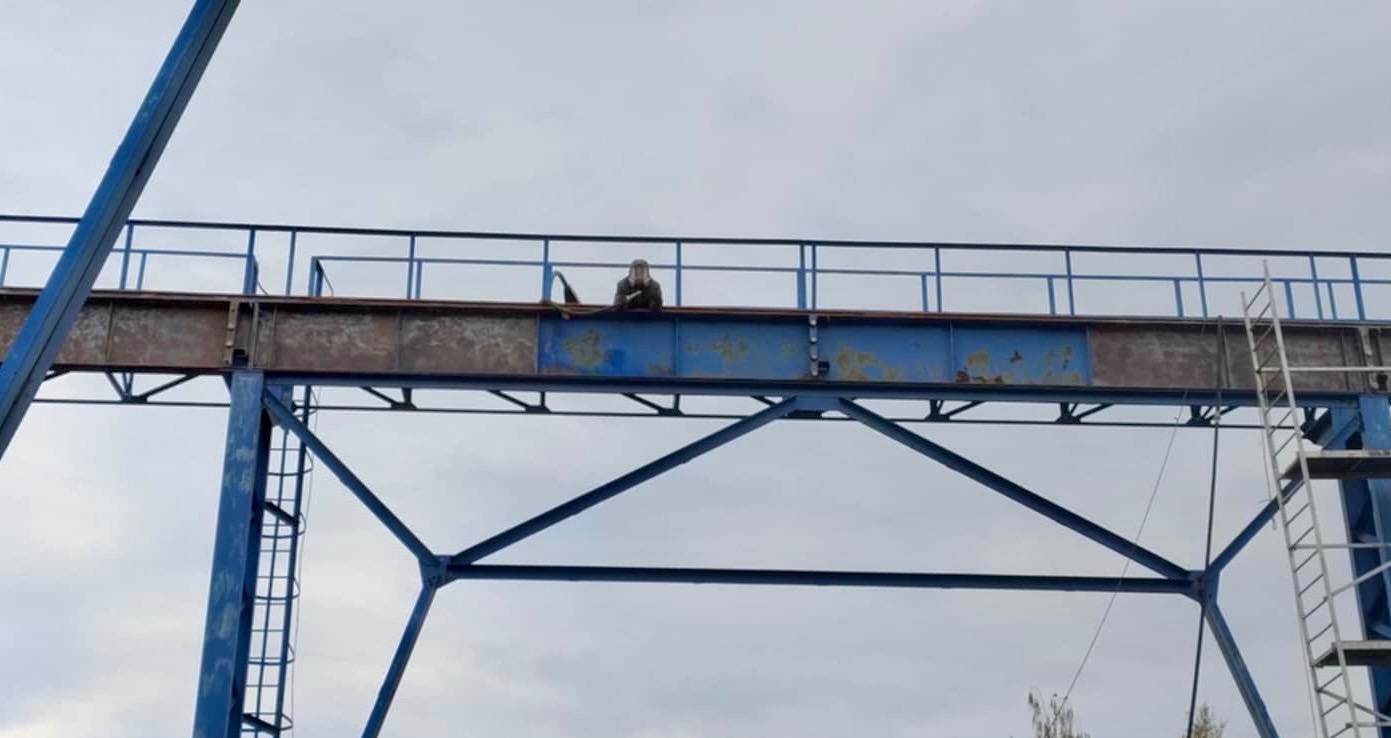Pracownik w kombinezonie na wysokości, na konstrukcji stalowej zardzewiałej w trakcie prac konserwacyjnych, widoczna drabina.