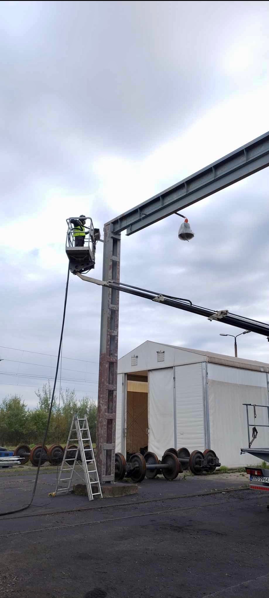 Pracownik w podnośniku koszowym czyści metalową konstrukcję na zewnątrz, widoczne koła kolejowe, drabina i biały namiot w tle, pochmurne niebo.