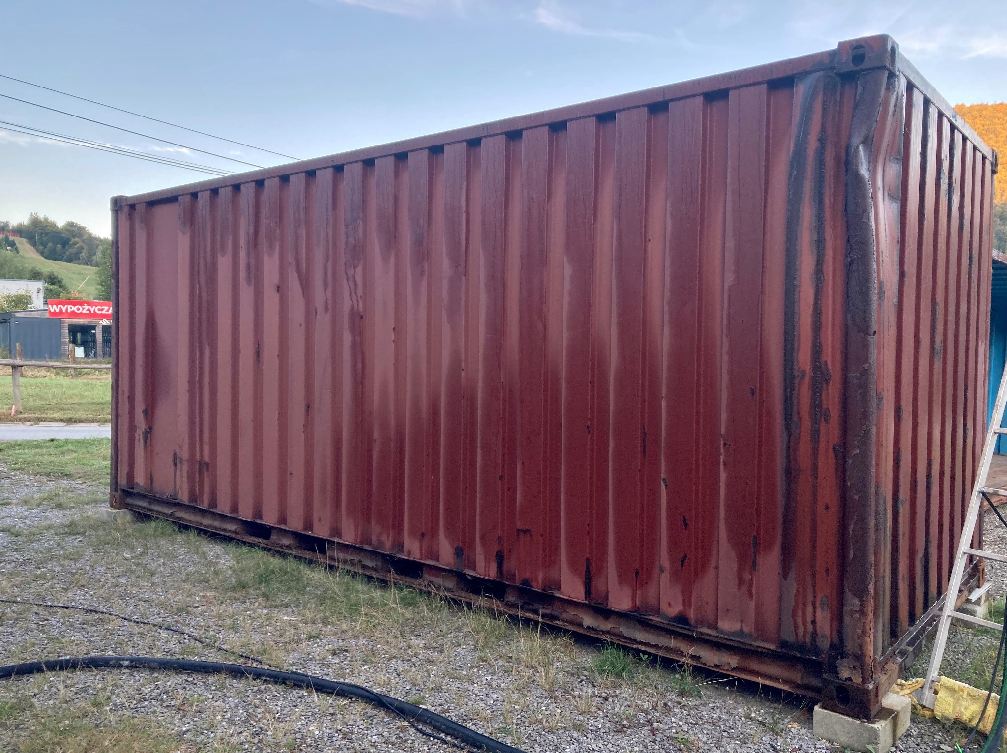 Czerwony, zardzewiały kontener transportowy z wgnieceniami na rogu, stojący na żwirowym podłożu, widoczna drabina oparta o ścianę kontenera, w tle budynek z napisem 'WYPOŻYCZAJ'.