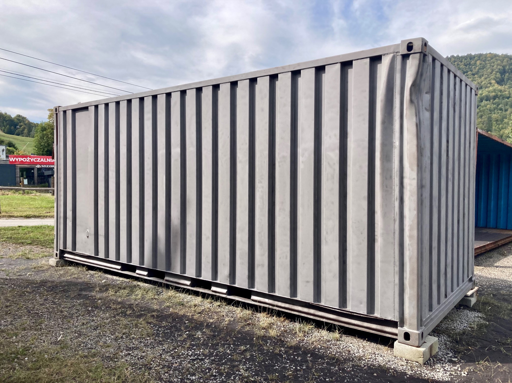 Szary kontener magazynowy ustawiony na betonowych bloczkach na żwirowym podłożu, z widocznym uszkodzeniem na jednej ze ścian, w tle zieleń drzew i błękitne niebo.