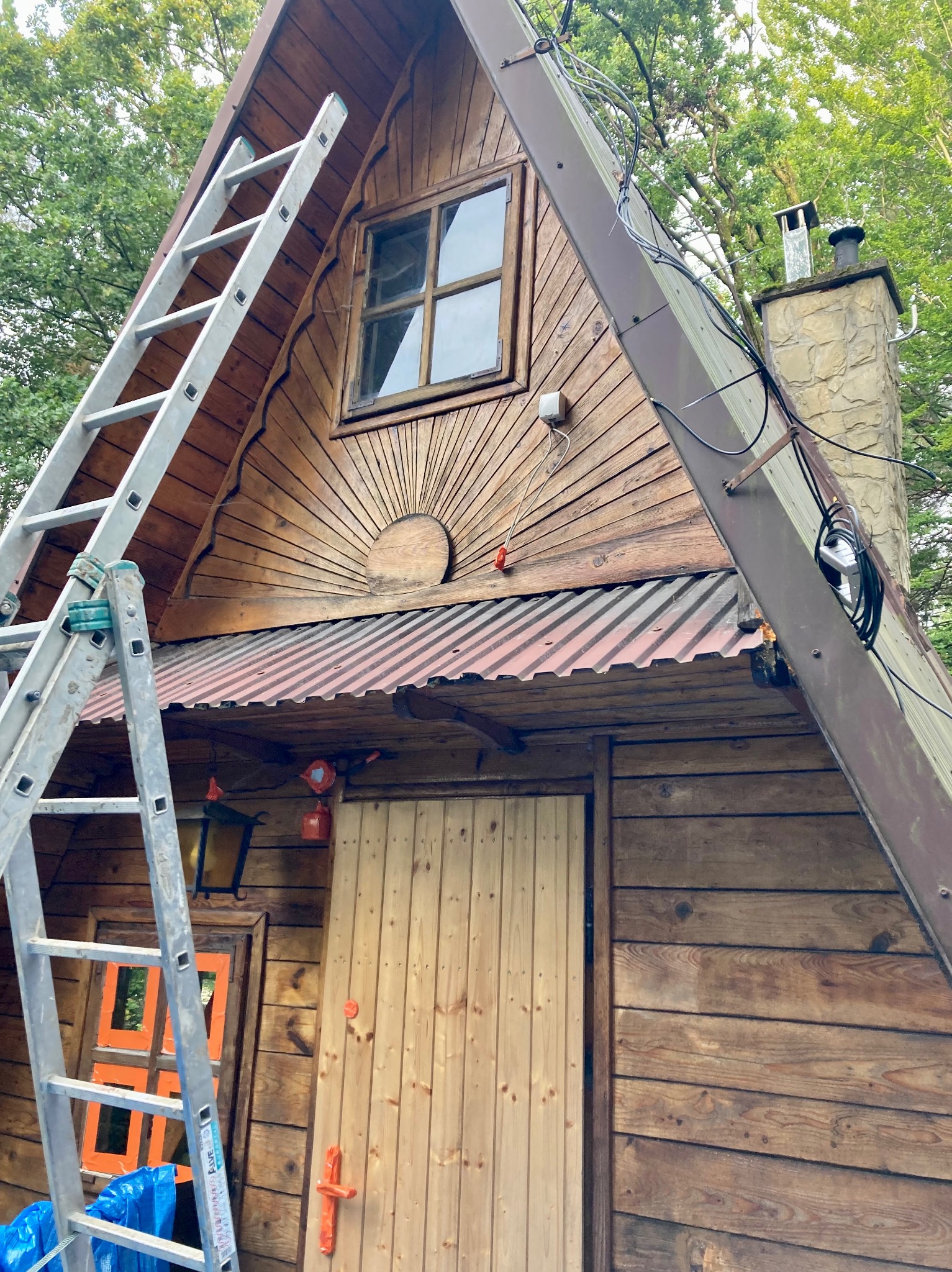 Drewniany domek w kształcie litery A z metalową drabiną opartą o ścianę, widoczny fragment dachu z kominem, okno z drewnianą ramą i ozdobny element w kształcie promieni słonecznych nad wejściem.