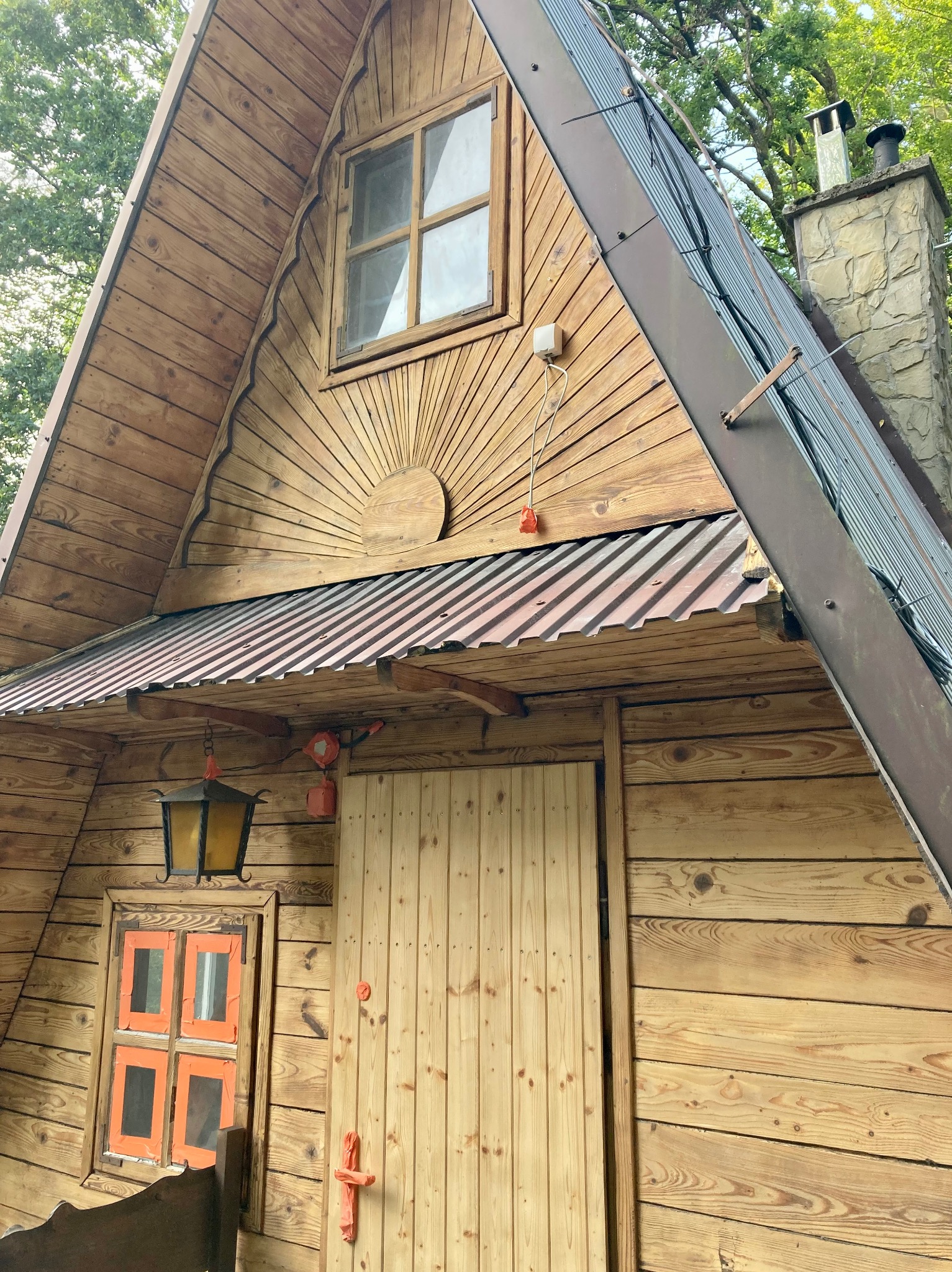 Drewniany domek letniskowy typu A-frame z widocznym dachem z blachy trapezowej, kamiennym kominem i dekoracyjnymi elementami drewnianymi wokół okien i drzwi, w otoczeniu zieleni.