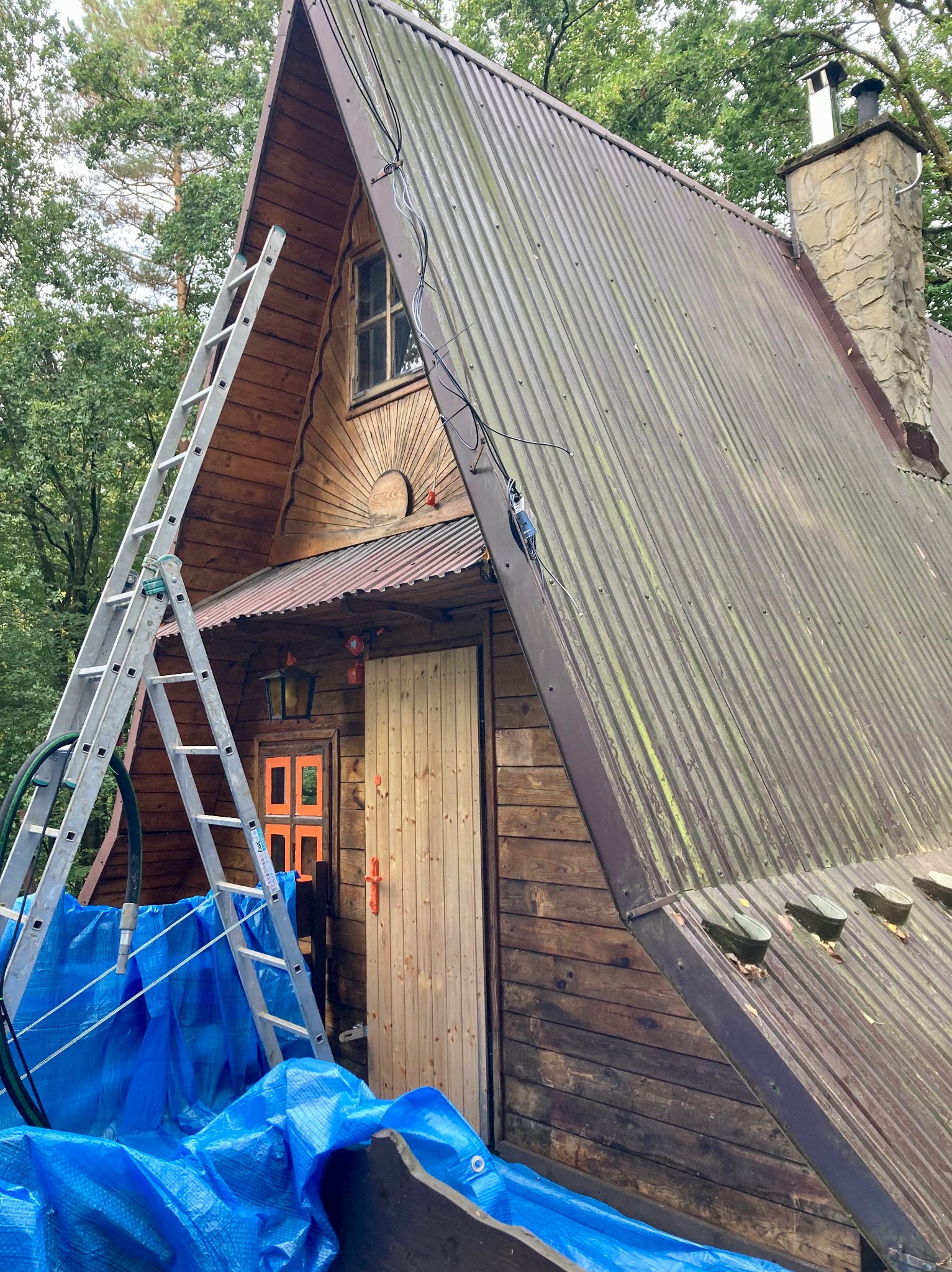 Drewniany domek typu A z metalowym dachem pokrytym mchem, drabina oparta o ścianę, zabezpieczenie niebieską plandeką, widoczny komin z kamienia.