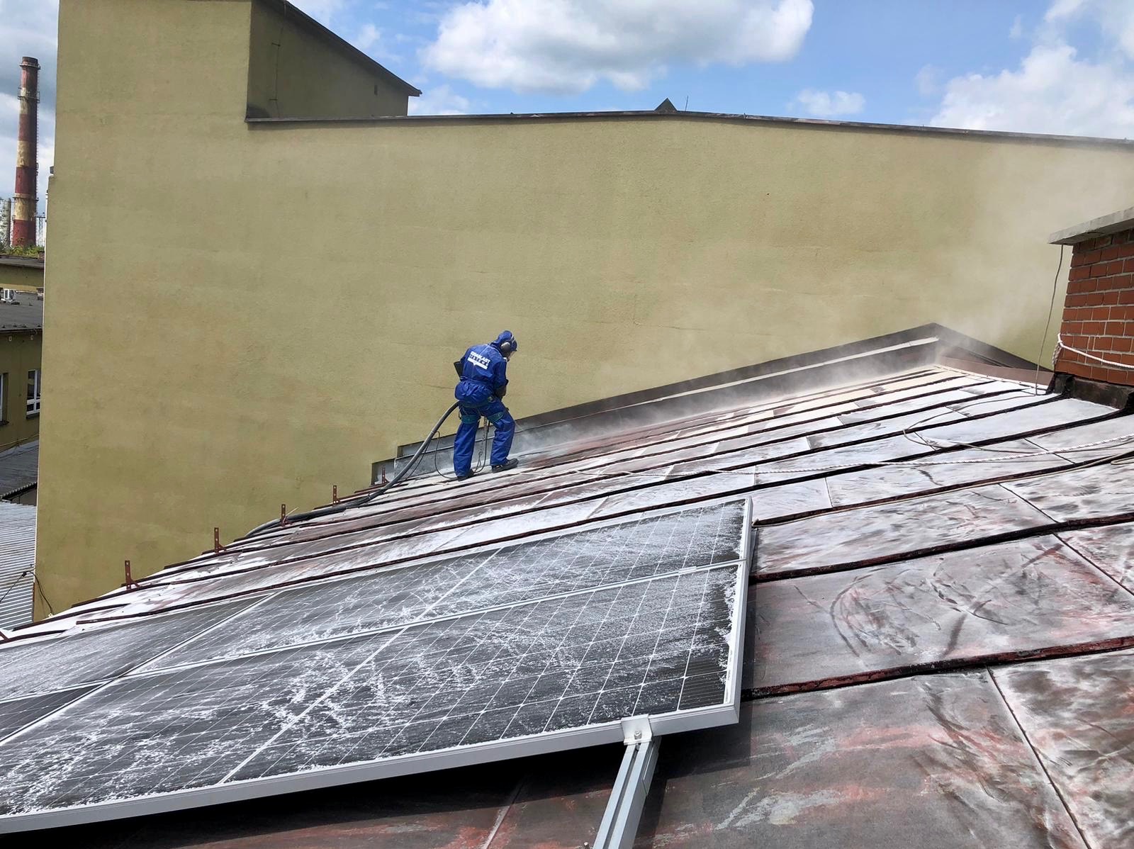 Pracownik w niebieskim kombinezonie ochronnym czyści dach z paneli słonecznych za pomocą węża pod ciśnieniem, budynek przemysłowy w tle.