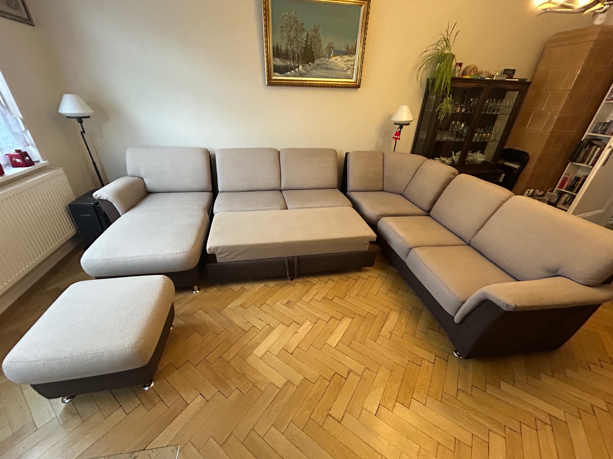 Duży, beżowo-brązowy narożnik z pufą na parkiecie w salonie, widok z góry, na ścianie obraz w złotej ramie.