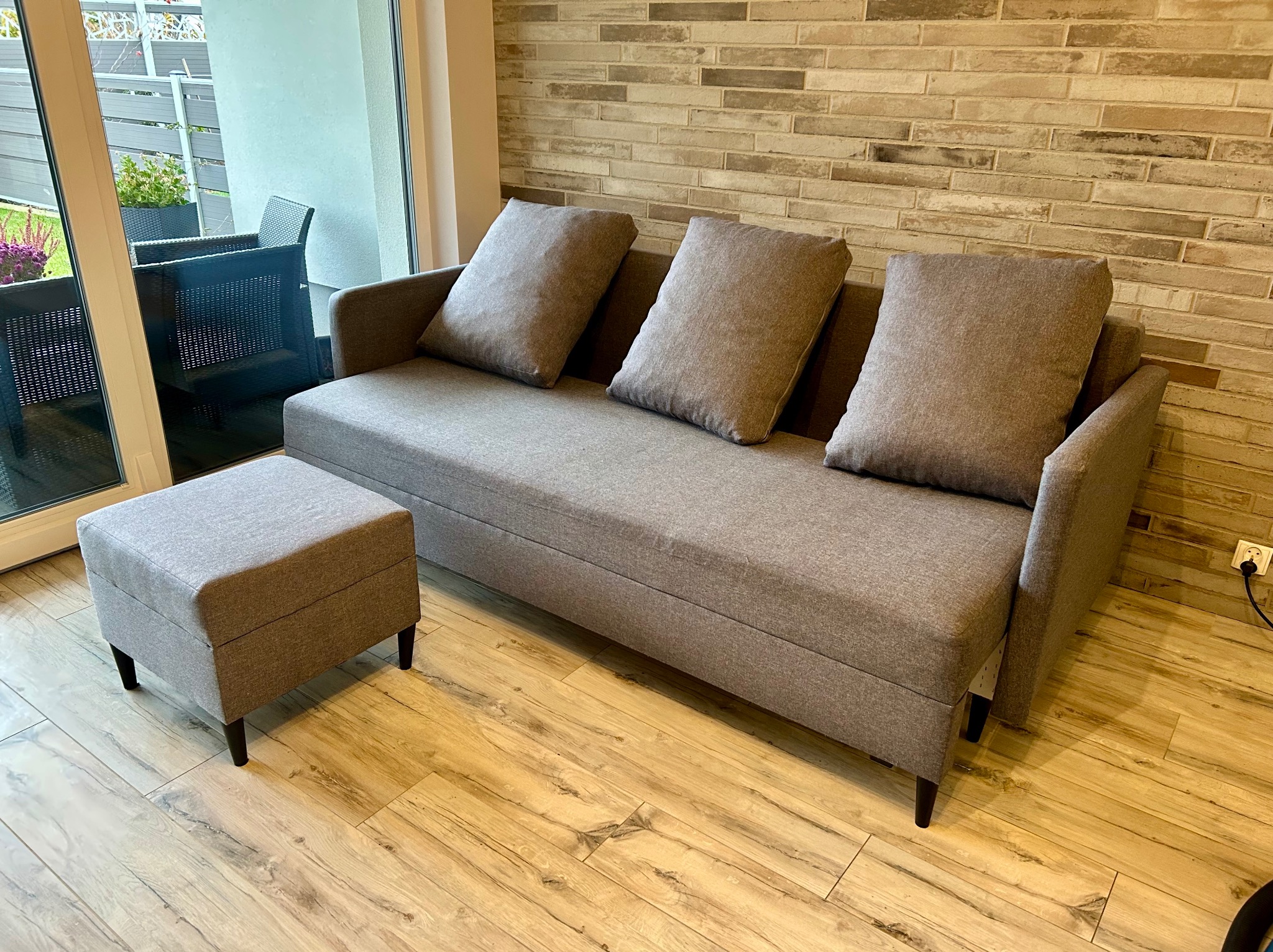 Nowoczesna szara sofa z trzema poduszkami i pufem na drewnianej podłodze, z widokiem na balkon przez przesuwne drzwi.