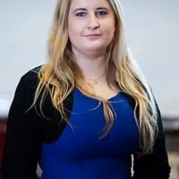 Portret kobiety o jasnych włosach, ubranej w niebieską sukienkę i czarny kardigan, z delikatnym naszyjnikiem. Uśmiech na twarzy, neutralne tło.