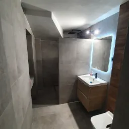 Nowoczesna łazienka z szarymi płytkami, podświetlanym lustrem nad drewnianą szafką umywalkową i wiszącą toaletą, oświetlona punktowymi lampami.