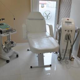 Yasumi Instytut Zdrowia i Urody - Białe, skórzane, regulowane krzesło zabiegowe w sterylnym, jasnym pomieszczeniu z urządzeniami do zabiegów kosmetycznych, w tle okno z naklejonym logo firmy.