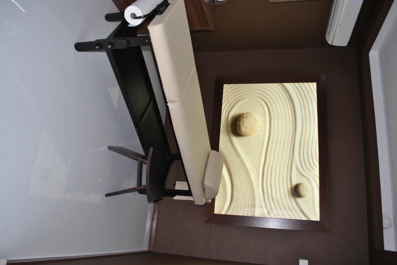 Beżowy fotel do zabiegów kosmetycznych z podkładem papierowym, ustawiony obok obrazu zen z piaskiem i kamieniami w ciemnej ramie.