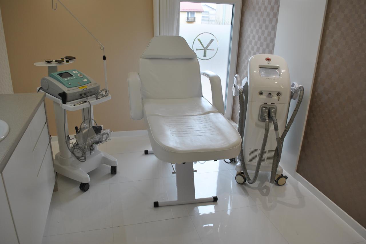 Białe, skórzane, regulowane krzesło zabiegowe w sterylnym, jasnym pomieszczeniu z urządzeniami do zabiegów kosmetycznych, w tle okno z naklejonym logo firmy.