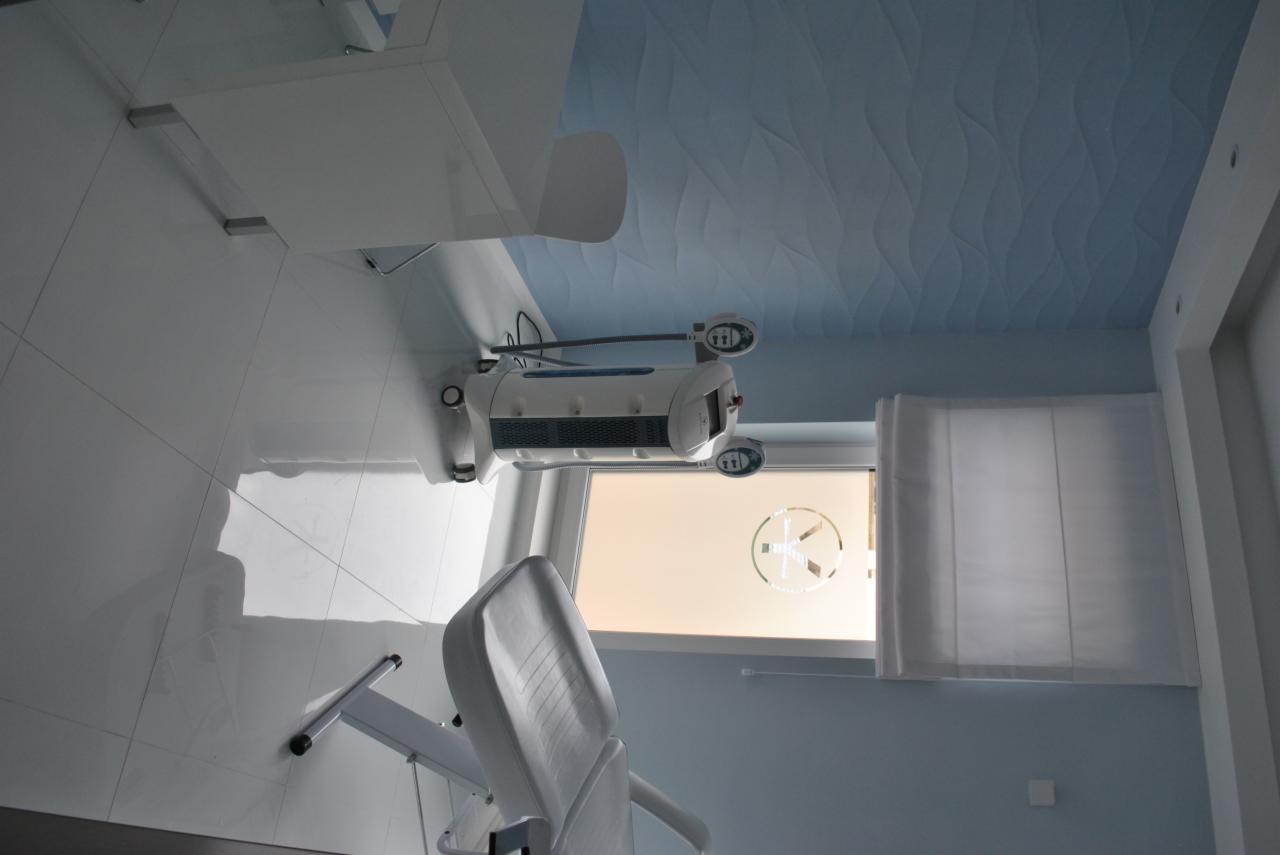 Jasne wnętrze gabinetu kosmetycznego z fotelem zabiegowym, lampą i oknem z naklejką logo.