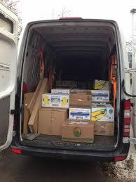 Wnętrze busa załadowane kartonami różnej wielkości, zabezpieczonymi pomarańczowymi pasami. Widoczne kartony z bananami i inne, częściowo zakryte paczki.