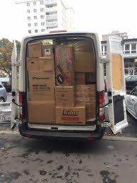 Wnętrze furgonetki załadowane kartonami różnej wielkości, widoczne etykiety firmowe na kartonach, otwarte tylne drzwi, w tle budynek mieszkalny i zaparkowane samochody.