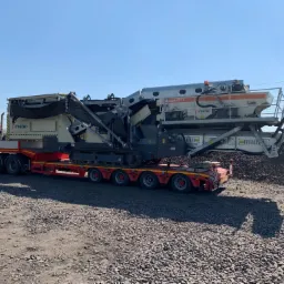 Ciężarówka transportuje mobilny przesiewacz kruszywa Metso na naczepie niskopodwoziowej, na tle niebieskiego nieba i hałdy kruszywa. Widoczne detale konstrukcyjne maszyny i ciągnika siodłowego.