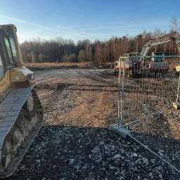 Żółty buldożer gąsienicowy z przodu, w tle teren po niwelacji z dwoma koparkami i zabezpieczeniem siatką, pod błękitnym niebem.