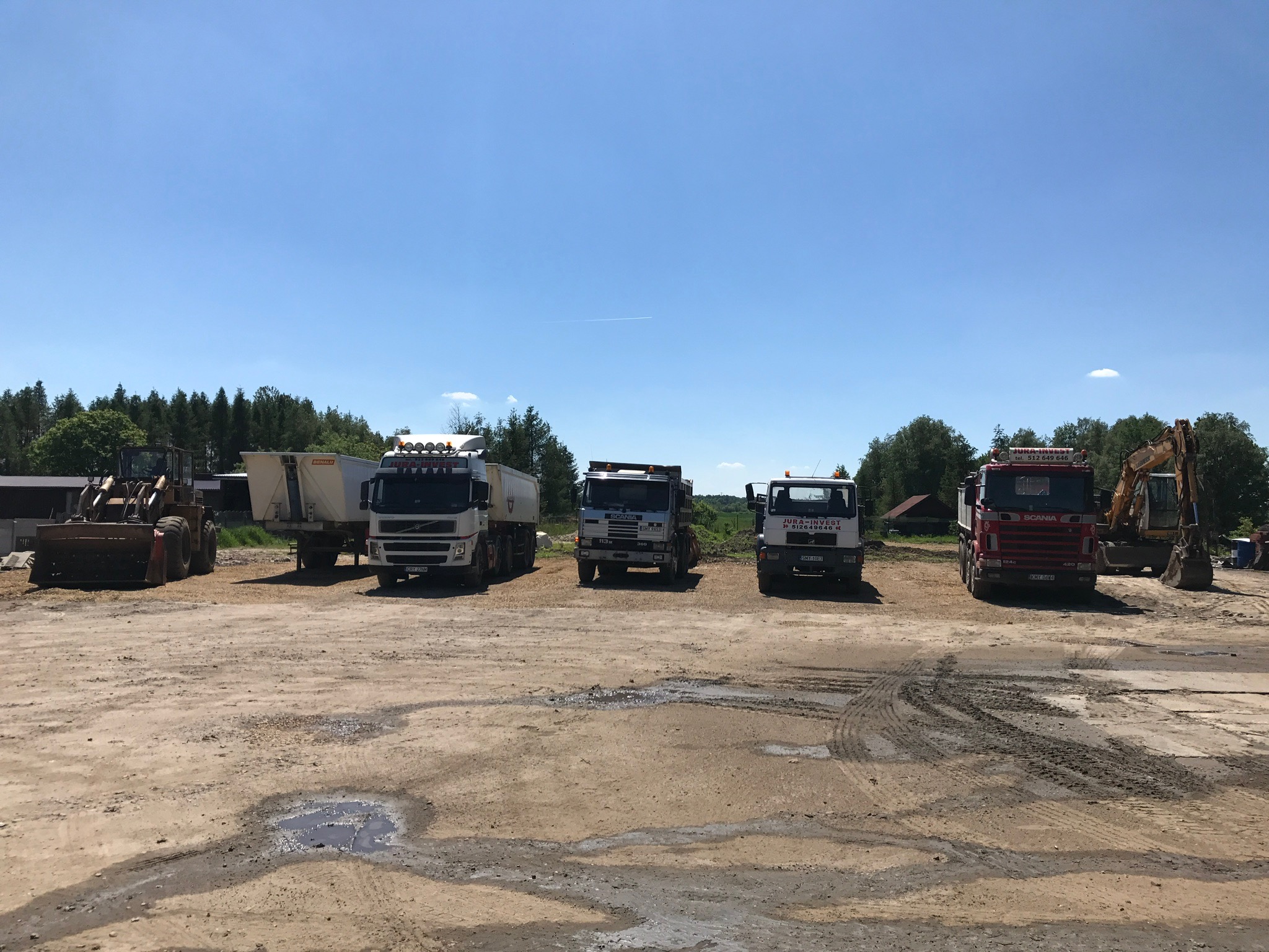 Sprzęt budowlany: ładowarka, wywrotki i koparka zaparkowane na placu budowy z błotnistym podłożem i kałużami, widoczne opony i ślady pojazdów na ziemi, w tle drzewa i błękitne niebo.