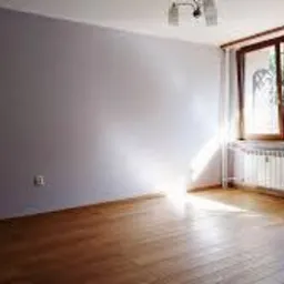 Pusty pokój z drewnianą podłogą, świeżo pomalowanymi ścianami w odcieniach szarości, kaloryferem pod oknem i wyraźnym światłem słonecznym wpadającym do wnętrza.