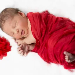 Śpiący noworodek owinięty w czerwoną tkaninę, obok czerwony kwiat na białym tle.