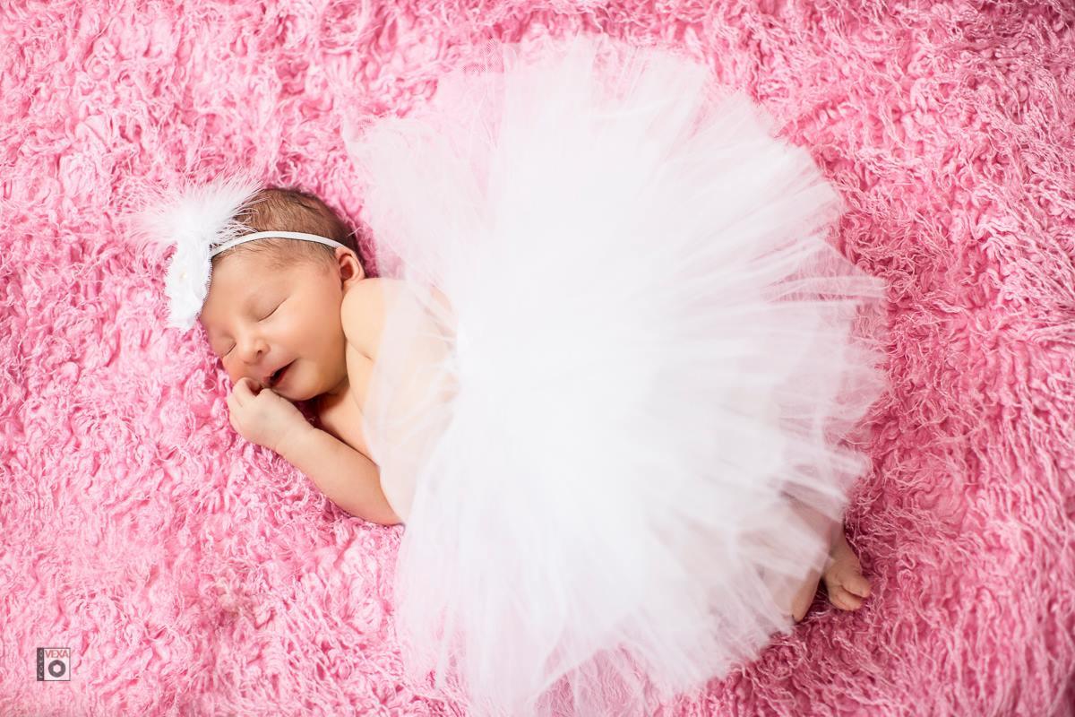 Śpiąca noworodka w białej tiulowej spódniczce i opasce z piórkiem na różowym, puszystym tle.