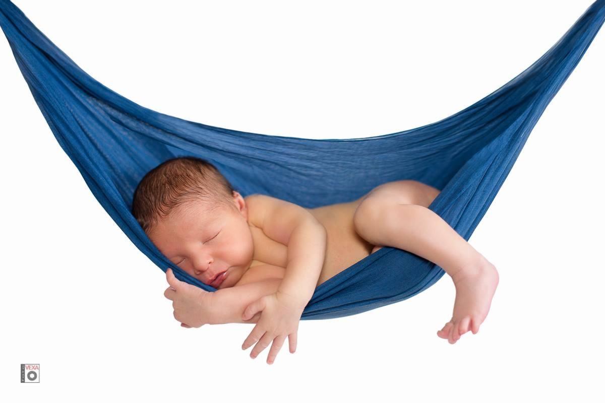 Noworodek śpiący w niebieskim hamaku na białym tle, delikatne oświetlenie, widoczne drobne rączki i stopy.