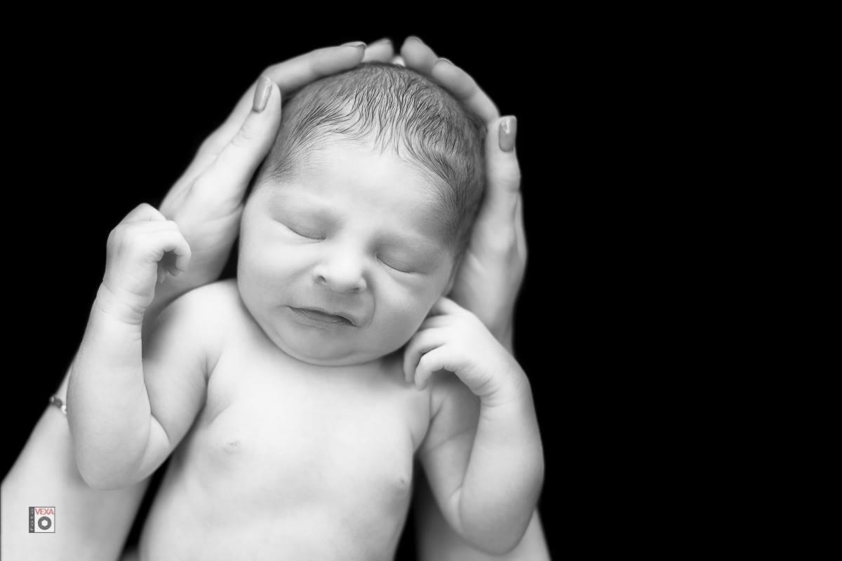 Monochromatyczne zbliżenie śpiącego noworodka, którego głowę i ramiona obejmują dłonie dorosłej osoby z delikatnym manicure, na jednolitym czarnym tle.