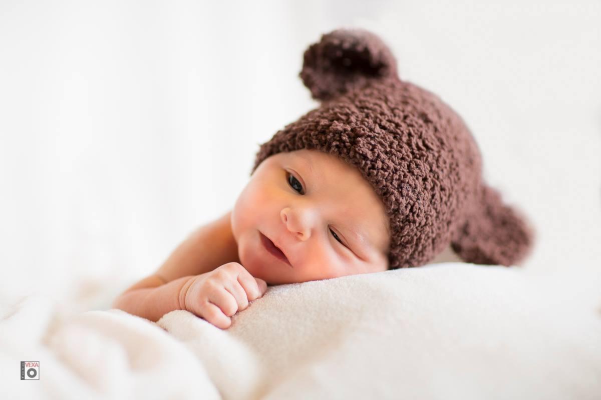 Noworodek w brązowej, dzierganej czapce z uszkami, leżący na białym kocyku i mrużący jedno oko.