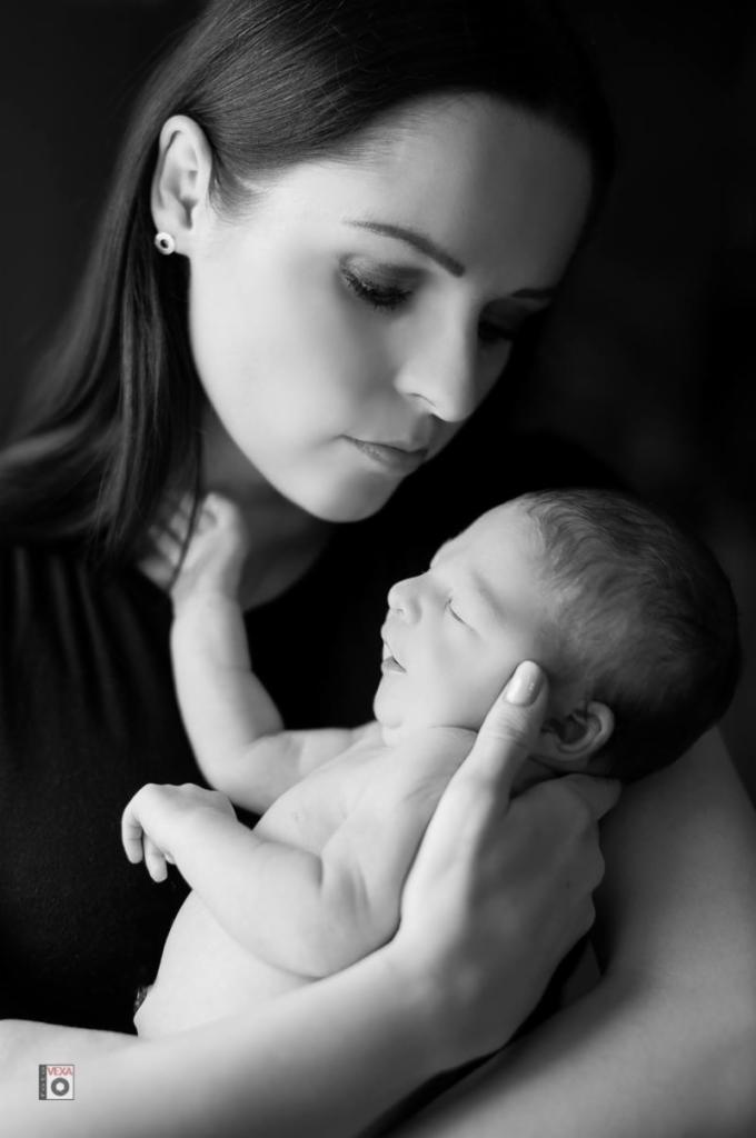 Czarno-białe zdjęcie matki spoglądającej z czułością na śpiącego noworodka, którego trzyma w ramionach, delikatnie podtrzymując jego główkę; widoczny mały kolczyk w uchu kobiety.
