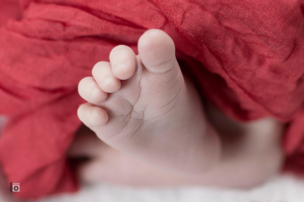 Makro zbliżenie na drobną stopę noworodka, z wyraźnie widocznymi żyłkami i fałdkami skóry, częściowo otuloną czerwoną, miękką tkaniną.